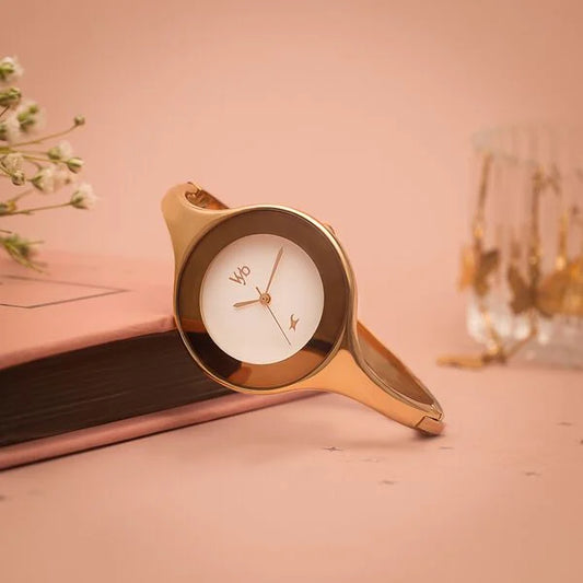 A rose gold-colored analog watch with a white dial, branded 'VYB FASTRACK', displayed on a pink surface with a floral design in the background.